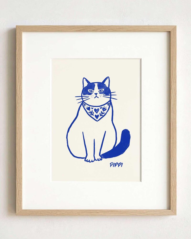 A framed art print featuring a blue ink illustration of a cat wearing a patterned bandana with hearts and floral motifs. The cat is sitting upright, and the name PIPPI is printed in blue ink below the cat on a cream background.