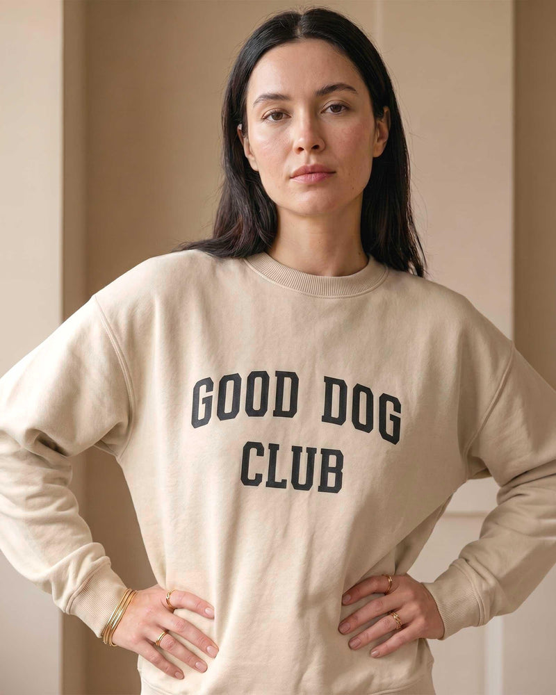 Woman wearing a sand-colored crewneck sweatshirt with bold black collegiate text reading Good Dog Club on the chest, styled simply against a warm neutral background.