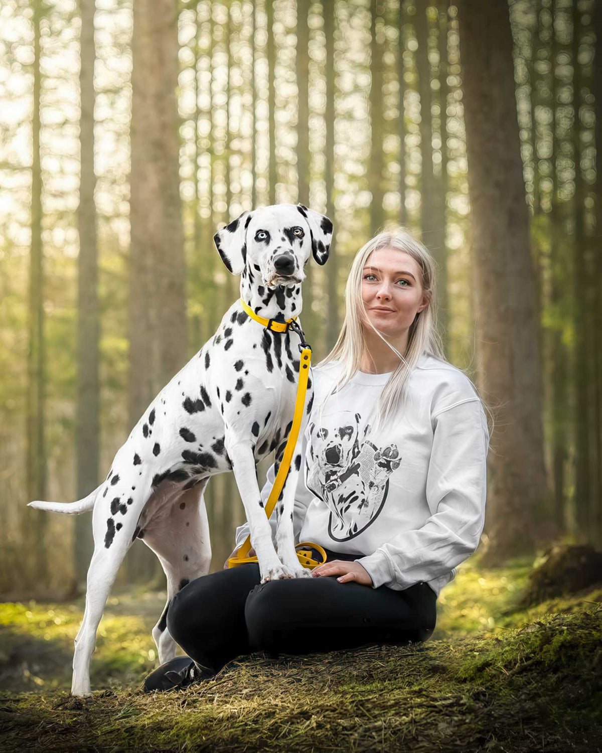 A woman sitting on a mossy log in a sunlit forest with a Dalmatian dog beside her. The woman wears a white sweatshirt with a black floral graphic and black pants, and the dog wears a yellow collar and leash.