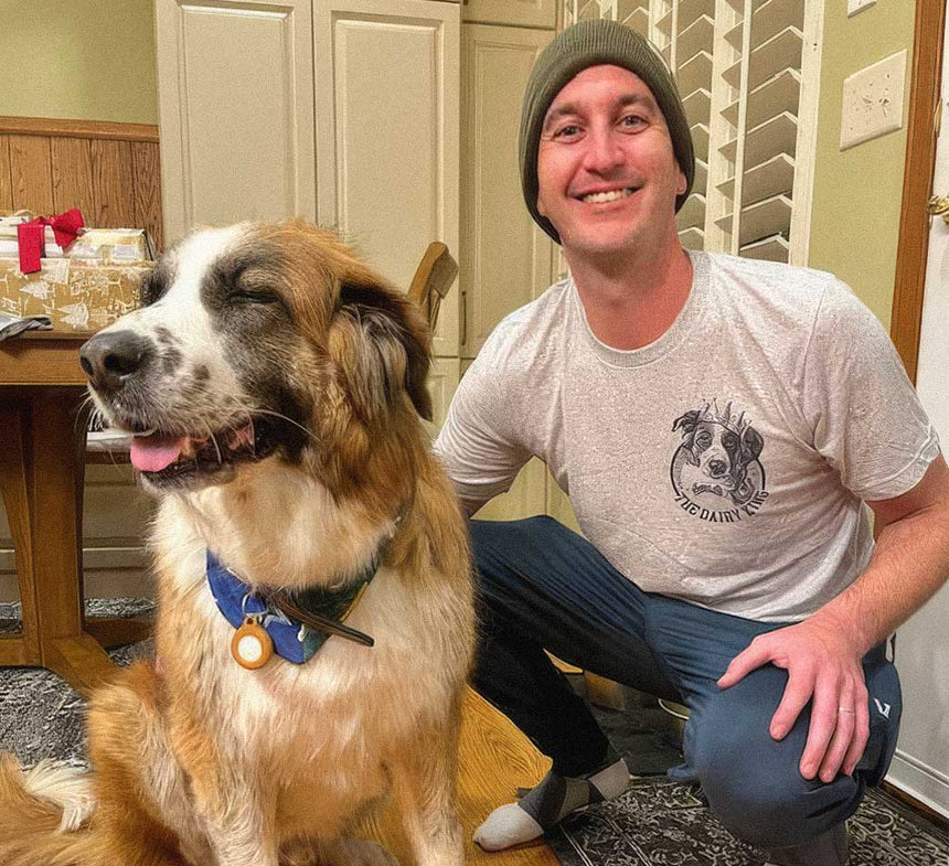 A smiling man wearing a gray beanie and a light gray t-shirt with a dog graphic, crouching next to a large brown and white dog wearing a blue collar in a home setting.