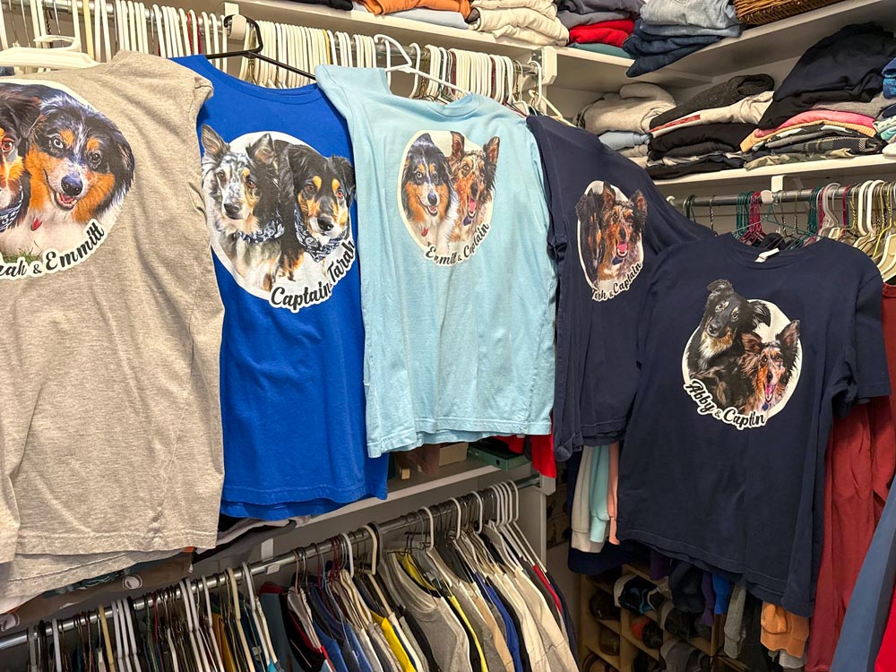 A row of custom graphic t-shirts featuring dog portraits, displayed on hangers in a closet or storage area. The shirts are shown in multiple colors including heather gray, royal blue, light blue, dark blue, and navy, each with a different dog design and text like 'Bake & Emmi' and 'Captain Chaos'.