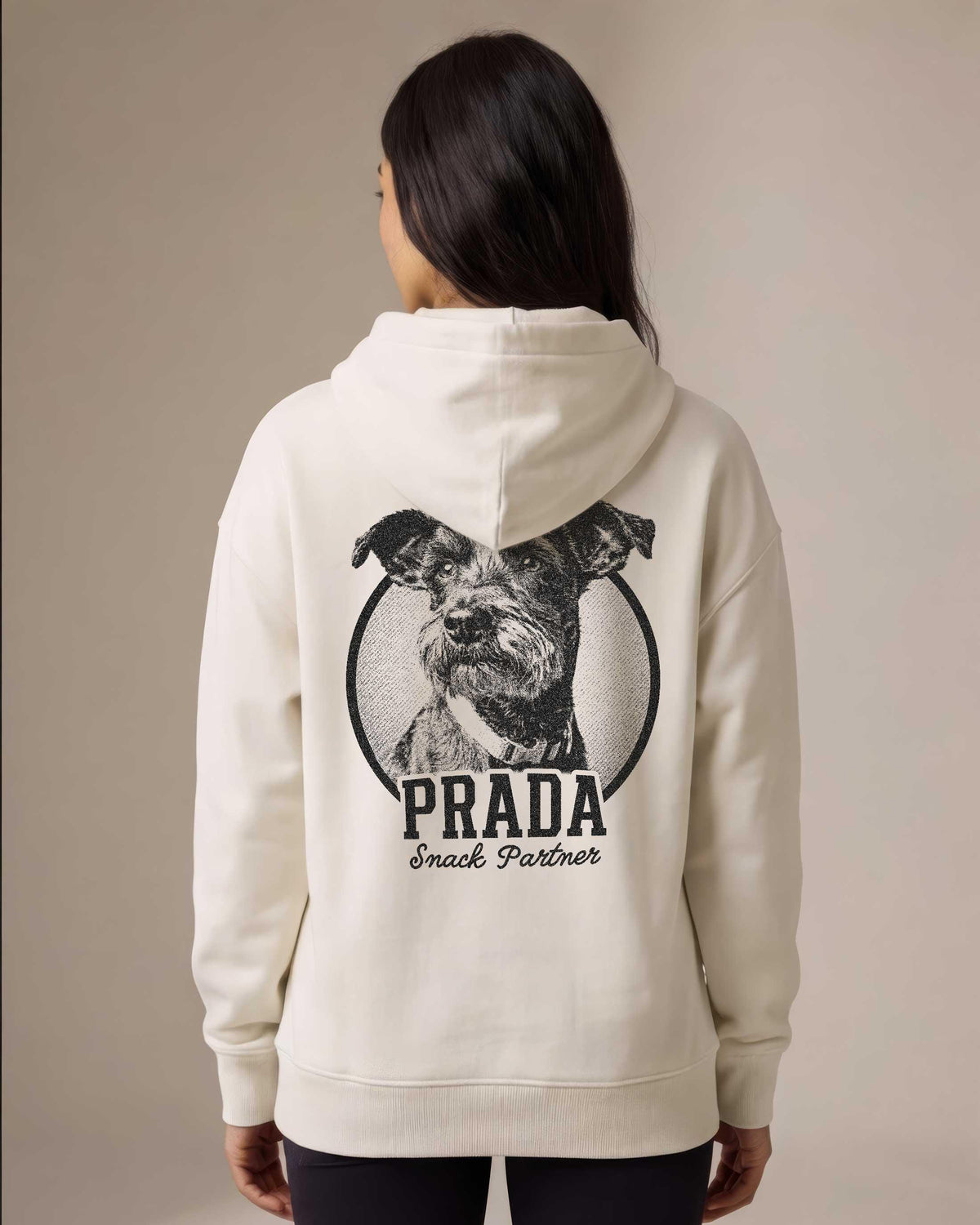 Woman wearing a cream pullover hoodie with a large custom dog portrait printed on the back inside a circular badge, showing a schnauzer terrier mix with the name Prada and tagline Snack Partner in bold vintage lettering.