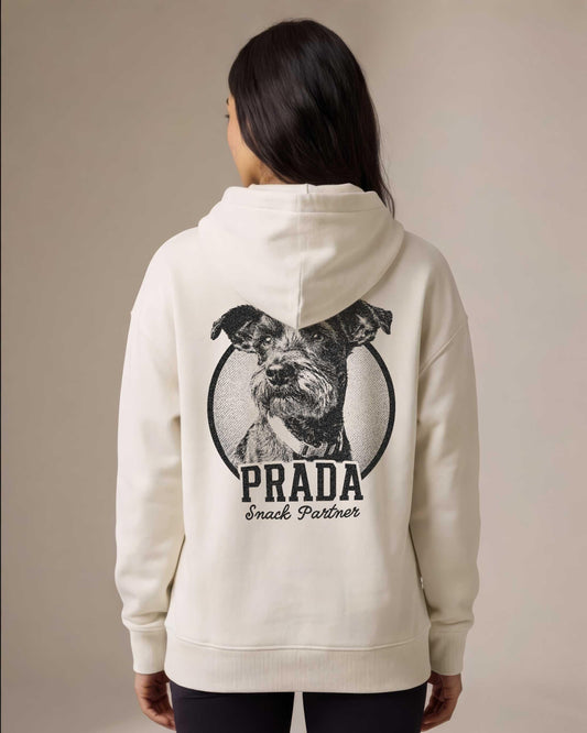 Woman wearing a cream pullover hoodie with a large custom dog portrait printed on the back inside a circular badge, showing a schnauzer terrier mix with the name Prada and tagline Snack Partner in bold vintage lettering.