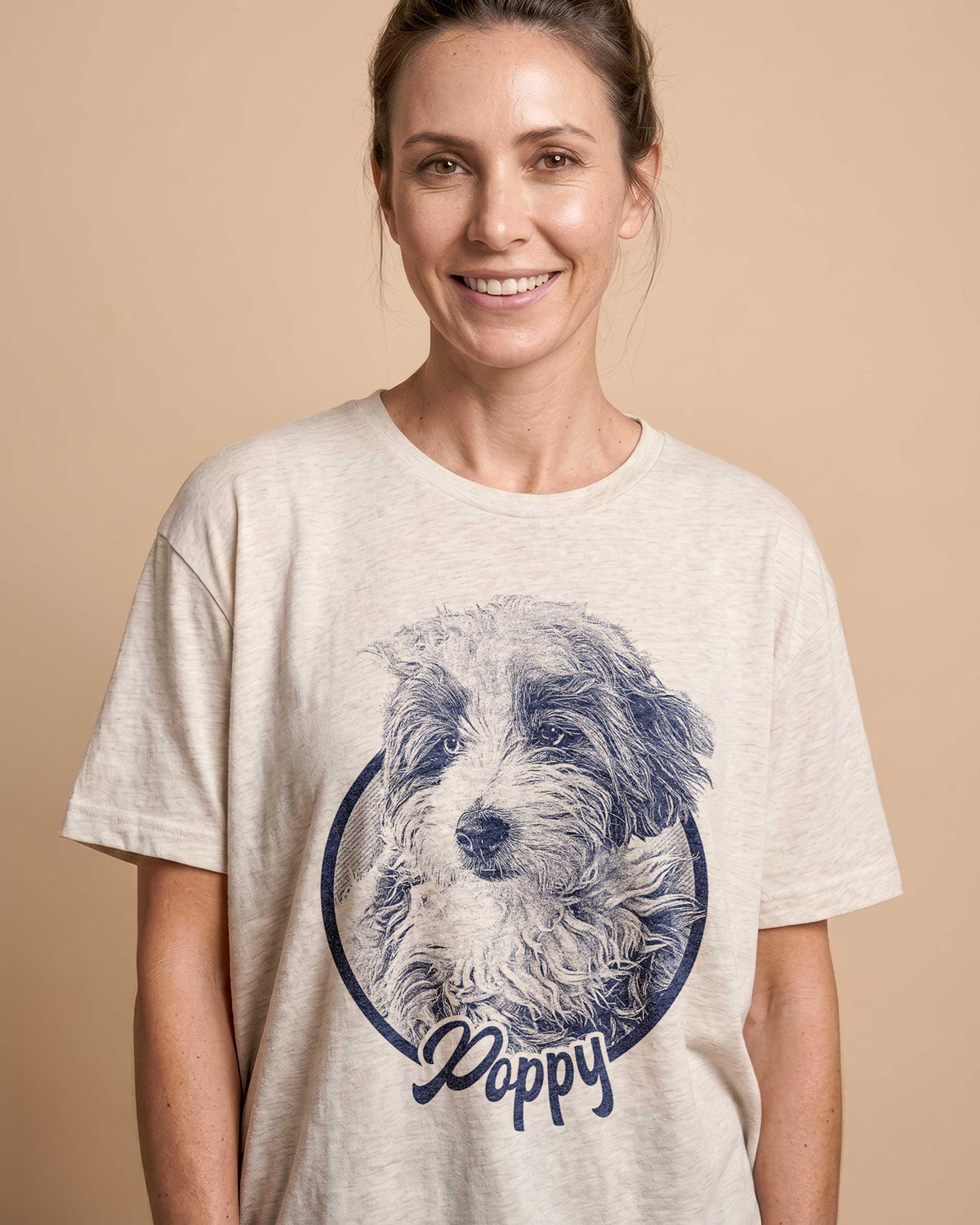 Smiling woman wearing a beige custom pet T-shirt featuring a detailed vintage blue illustration of a happy dog named “Chewy.” The design is minimalist and vintage-inspired, with a circular frame around the pet portrait. The soft natural lighting and neutral background highlight the cozy, personalized style  perfect for showcasing custom dog lover gifts or memorial keepsake shirts.