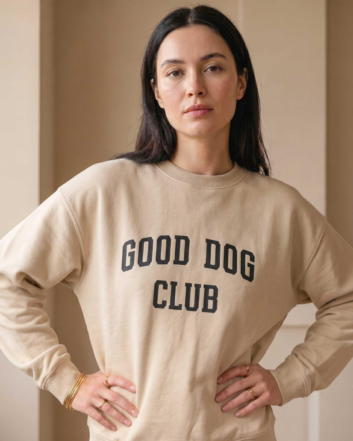 Woman wearing a sand-colored crewneck sweatshirt with bold black collegiate text reading Good Dog Club on the chest, styled simply against a warm neutral background.