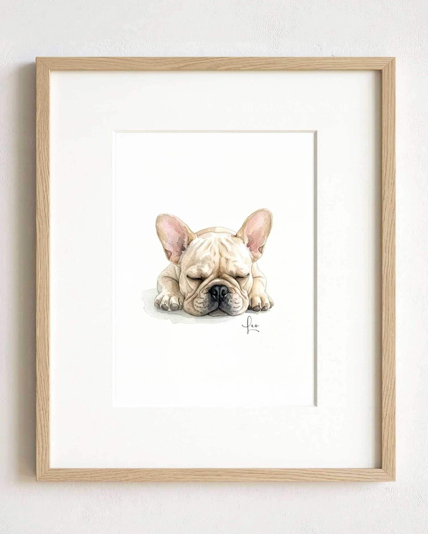 A framed watercolor illustration of a sleeping cream-colored French Bulldog with large ears and a black nose. The artwork is centered within a light-toned wooden frame, featuring a signature in the bottom right corner of the painting.