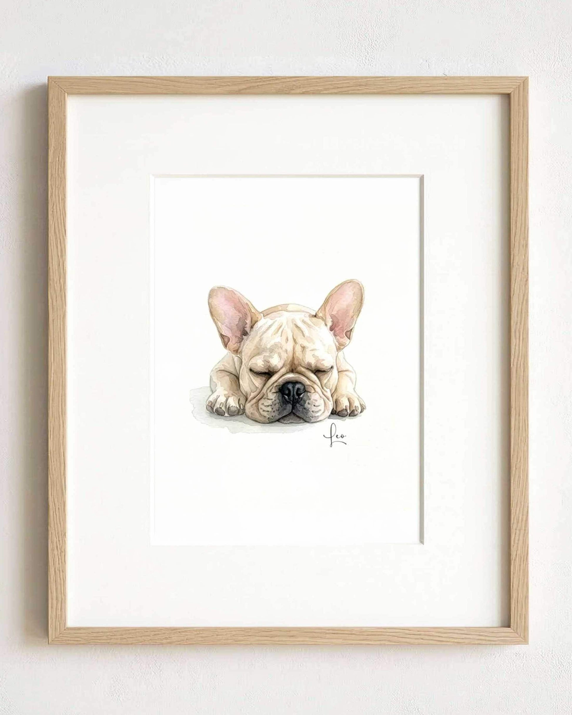 A framed watercolor illustration of a sleeping cream-colored French Bulldog with large ears and a black nose. The artwork is centered within a light-toned wooden frame, featuring a signature in the bottom right corner of the painting.