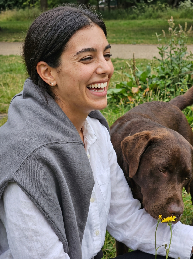 A woman smiling outdoors next to a brown dog, wearing a white shirt and a gray cardigan. The background shows a green park setting.